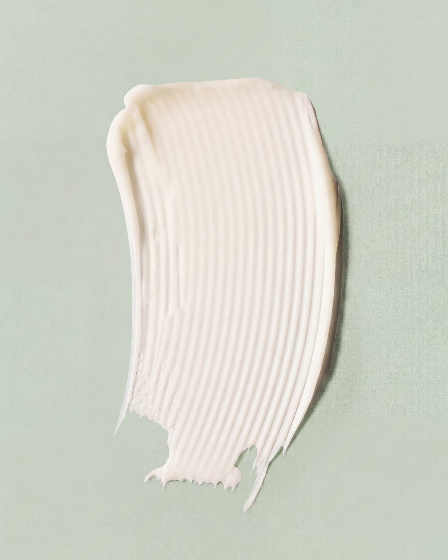 A textured smear of off-white cream or lotion with visible ridges, placed on a soft sage green background.
